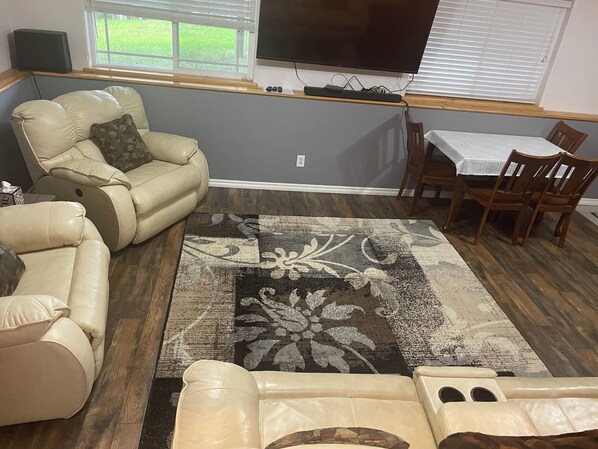 A living room with a beige leather sofa, a patterned rug, a wooden dining table with chairs, and a flat-screen TV mounted on the wall.