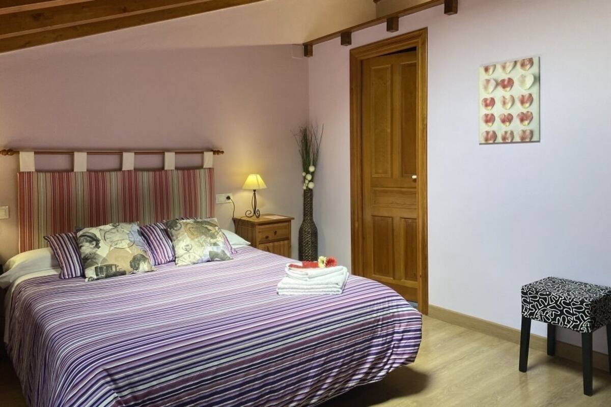 Un dormitori amb un llit, una taula de nit, una làmpada, una porta i un petit tamboret.