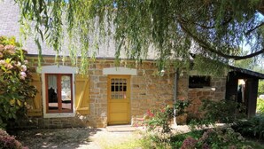 A stone cottage with a yellow door, a small window with curtains, and a garden with pink flowers.