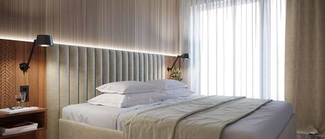A modern bedroom with a large bed, a bedside table, a lamp, and a window with sheer curtains.