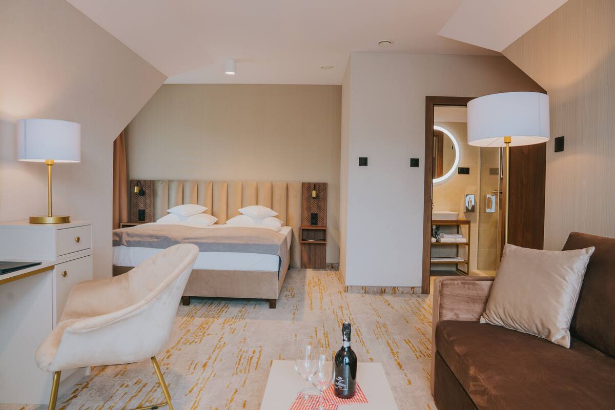 A hotel room with a bed, a sitting area with a chair and a sofa, a small table with a bottle and glass, and a mirror in the corner.