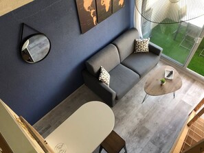 A modern living room with a grey sofa, a round mirror, a small round table with a plant, and a large window with a view of a green lawn.