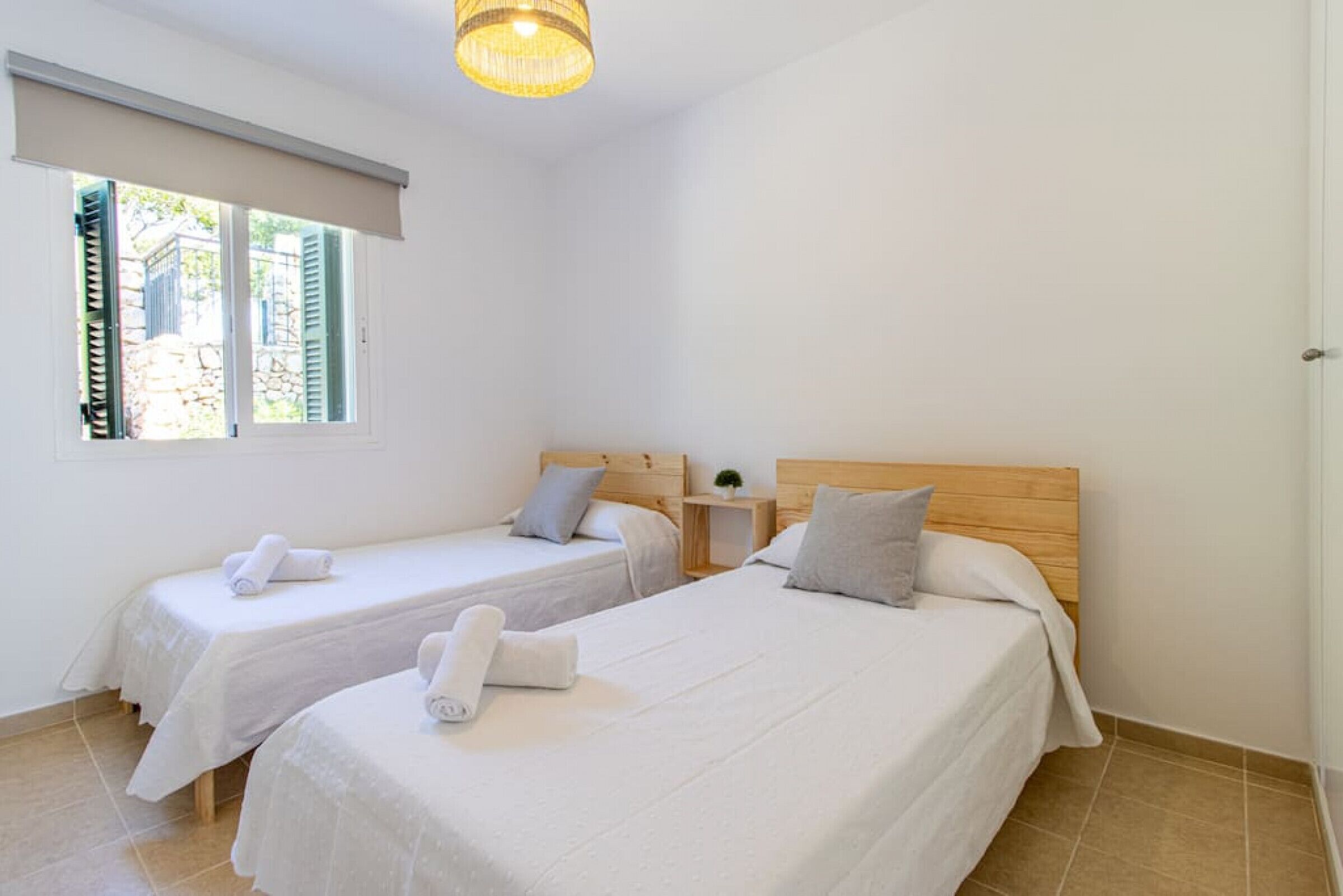 A hotel room with two single beds, a wooden headboard, a small bedside table with a lamp, and a window with shutters.