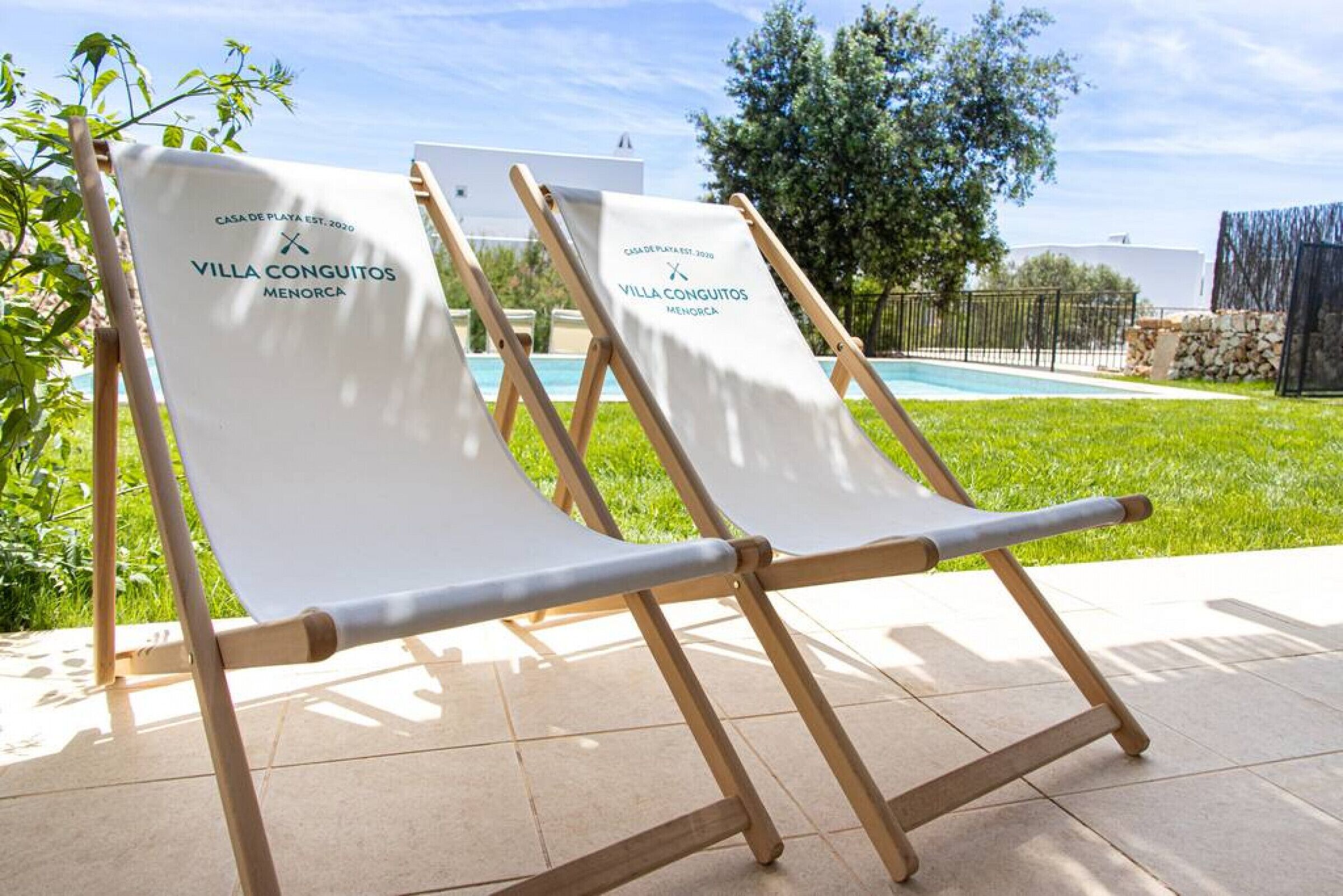 Two lounge chairs with a banner reading "VILLA CONQUITOS MENORCA" and a pool in the background.