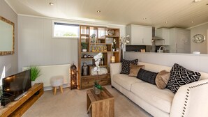 A living room with a sofa, a coffee table, a TV on a wooden stand, a bookshelf, and a kitchen in the background. - Inside No. 1 Retreat (Carnforth)