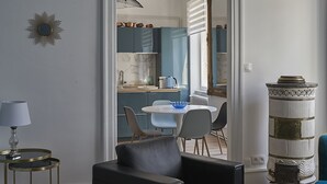 Un salón moderno con un sofá de cuero negro, una mesa de centro redonda y un jarrón azul. Se ve un comedor con una mesa y sillas blancas a través de un vano abierto.