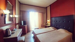 A hotel room with two beds, a desk with a kettle, a television, and a large window with curtains. - Park Hotel Volos (Volos)