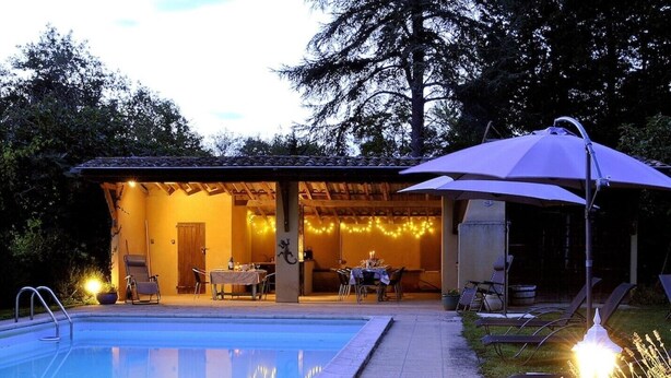 Un espace aménagé au bord de la piscine, avec un patio couvert, des sièges d’extérieur et une ambiance chaleureuse grâce à un éclairage soigné.