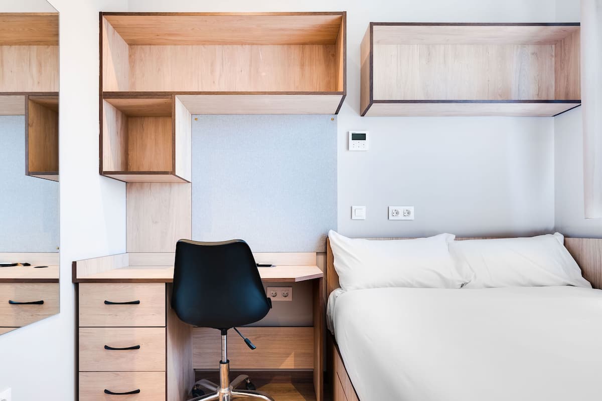 Ein Schlafzimmer mit einem Holztisch, einem schwarzen Bürostuhl, einem Bett mit weißer Bettwäsche und einem wandmontierten Lichtschalter.