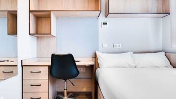 Quarto com escrivaninha de madeira, cadeira de escritório preta, cama com roupa de cama branca e interruptor de luz fixado na parede.