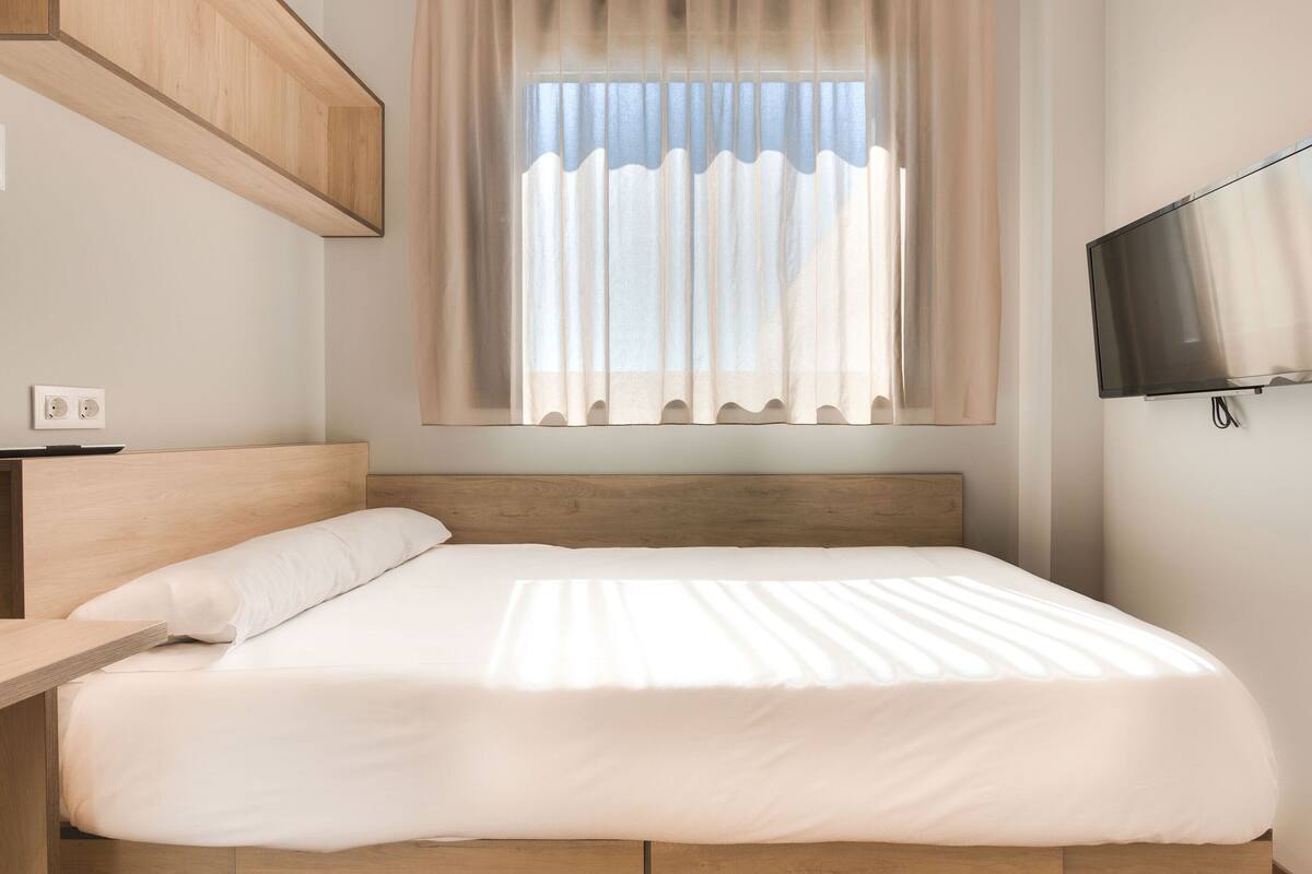 Ein modernes Schlafzimmer mit einem Holzkopfteil, einem Bett mit weißer Bettwäsche, einem Nachttisch, einem an der Wand montierten Fernseher und einem Fenster mit durchsichtigen Vorhängen.