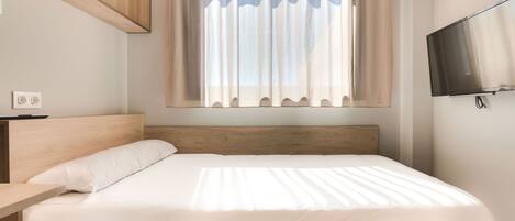 Une chambre moderne avec une tête de lit en bois, un lit recouvert de linge de lit blanc, une table de chevet, un téléviseur fixé au mur et une fenêtre ornée de rideaux légers.