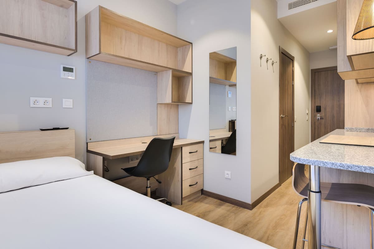 Ein modernes Hotelzimmer mit Bett, Schreibtisch, Stuhl, Spiegel und einer Küchenzeile.