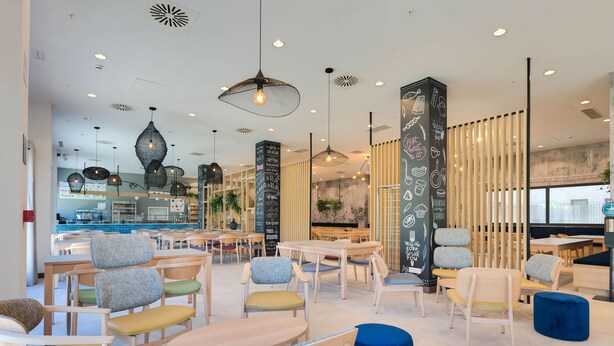 Interior de una cafetería moderna con mesas y sillas de madera, lámparas colgantes y una pared con menú en la pizarra.