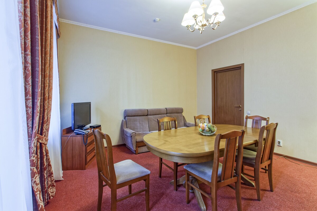A room with a round wooden dining table, six wooden chairs, a sofa, a television, and a door.
