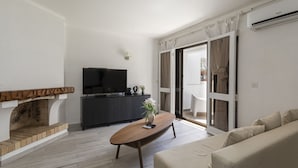 A modern living room with a fireplace, a flat-screen TV, a sofa, and a dining area visible through a glass door. - Vilamoura House Garden by Hopstays | Marina view (Vilamoura)