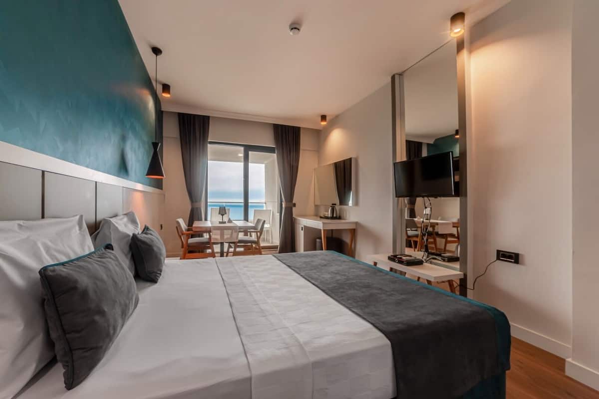 A modern hotel room with a large bed, a desk, a TV, and a view of the sea.