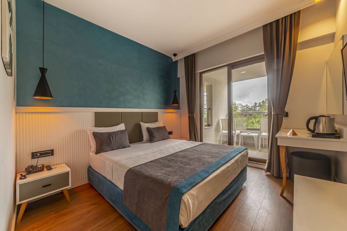 A modern hotel room with a large bed, bedside tables, a desk with a kettle, and a balcony with a view.