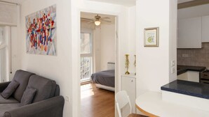 A modern living room with a gray sofa, a dining area with a white table and chairs, and a kitchen with white cabinets.