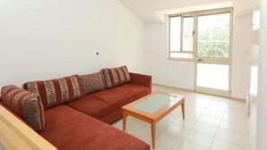 A living room with a red sofa, patterned cushions, a wooden coffee table, and a large window with a view of greenery. - Keren Kayemet - Four Bedroom Apartment, Sleeps 8 (Jerusalem)