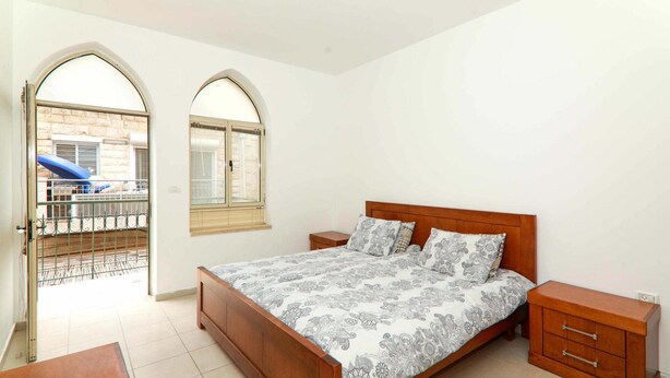 A bedroom with a wooden bed, two bedside tables, a wardrobe, and two arched windows. - Keren Kayemet - Four Bedroom Apartment, Sleeps 8 (Jerusalem)