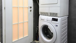 A laundry room with a stacked washer and dryer, a frosted glass door, and grey walls. - King David Elegance - Two Bedroom Apartment, Sleeps 4 (Jerusalem)