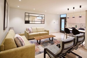 A modern living room with a sofa, coffee table, and chairs, a kitchen area with a dining table and chairs, and a large mirror on the wall.