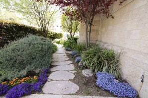 Un camí de pedra flanquejat per diferents plantes i flors, que condueix a un edifici amb una paret de pedra.