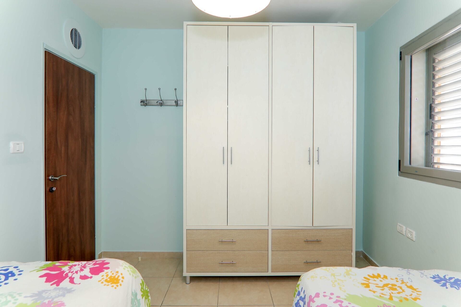 A bedroom with a wardrobe, two beds, a window, and a light switch.