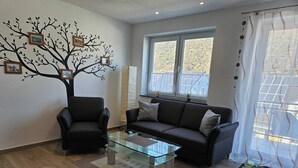 A living room with a glass coffee table, two sofas, a decorative tree wall, and a window with a view. - Ferienhaus Moselcharme "moselblick" (Alken)