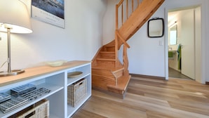Un escalier avec des rampes en bois menant à l’étage supérieur, un mur blanc orné d’un tableau encadré, une étagère blanche avec des paniers et des outils, et une lampe sur la gauche.