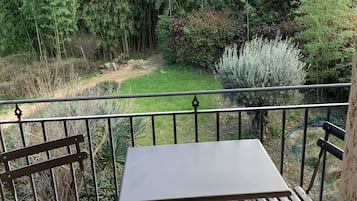 Ein Balkon mit Tisch und zwei Stühlen, mit Blick auf einen Garten mit verschiedenen Pflanzen und Bäumen.
