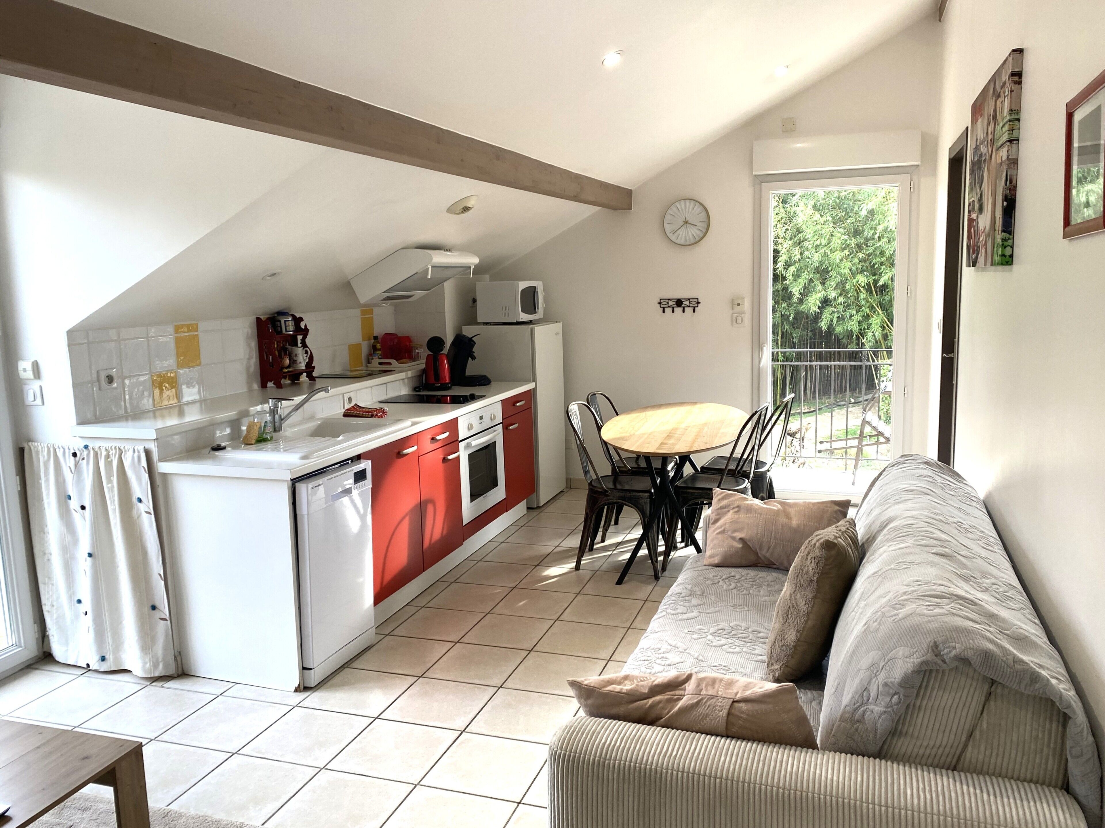A modern kitchen with red and white cabinetry, a dining area with a wooden table and chairs, and a living room with a sofa and a large window.