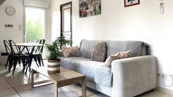 A modern living room with a sofa, a coffee table, and a dining area with chairs. There are two paintings on the wall, a clock, and a power outlet.