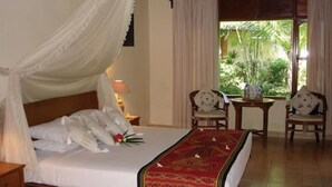 A bedroom with a canopy bed, a red decorative rug, two chairs, a small table, and a window with curtains. - Puri Benoa Suites Bungalows (Nusa Dua)