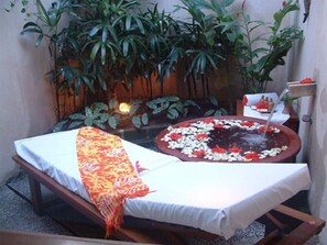 Un área de spa con una camilla de masaje, una bañera de hidromasaje con pétalos de flores y abundante vegetación al fondo.