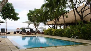 A swimming pool with lounge chairs and umbrellas, surrounded by trees and a view of the ocean. - Puri Benoa Suites Bungalows (Nusa Dua)