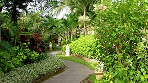 A curved pathway in a lush garden with various plants and a statue. - Puri Benoa Suites Bungalows (Nusa Dua)