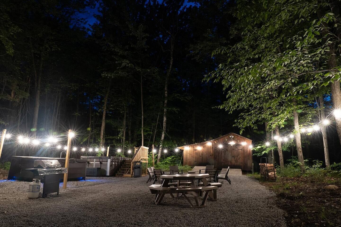 An outdoor patio area with picnic tables and a barbecue grill, surrounded by trees and a wooden building.
