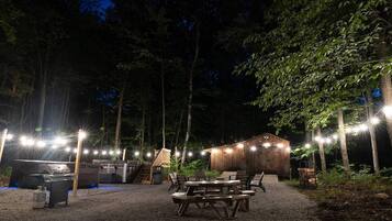 An outdoor patio area with picnic tables and a barbecue grill, surrounded by trees and a wooden building.