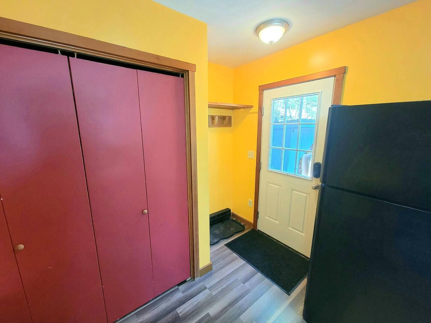 Un corridoio con una porta rossa chiusa, una porta bianca con una finestra e un frigorifero nero.