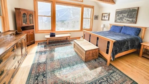 Une chambre avec une grande fenêtre, des poutres en bois, un lit, un banc, une armoire et un tapis.