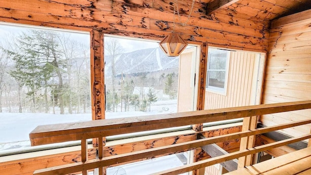 L’intérieur d’un chalet en bois, avec un escalier menant à un balcon, de grandes fenêtres et une vue sur une forêt enneigée.
