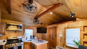 A wooden cabin interior with a kitchen featuring a stove, oven, and dishwasher, a dining area with a table and chairs, and a living area with a couch and a TV.