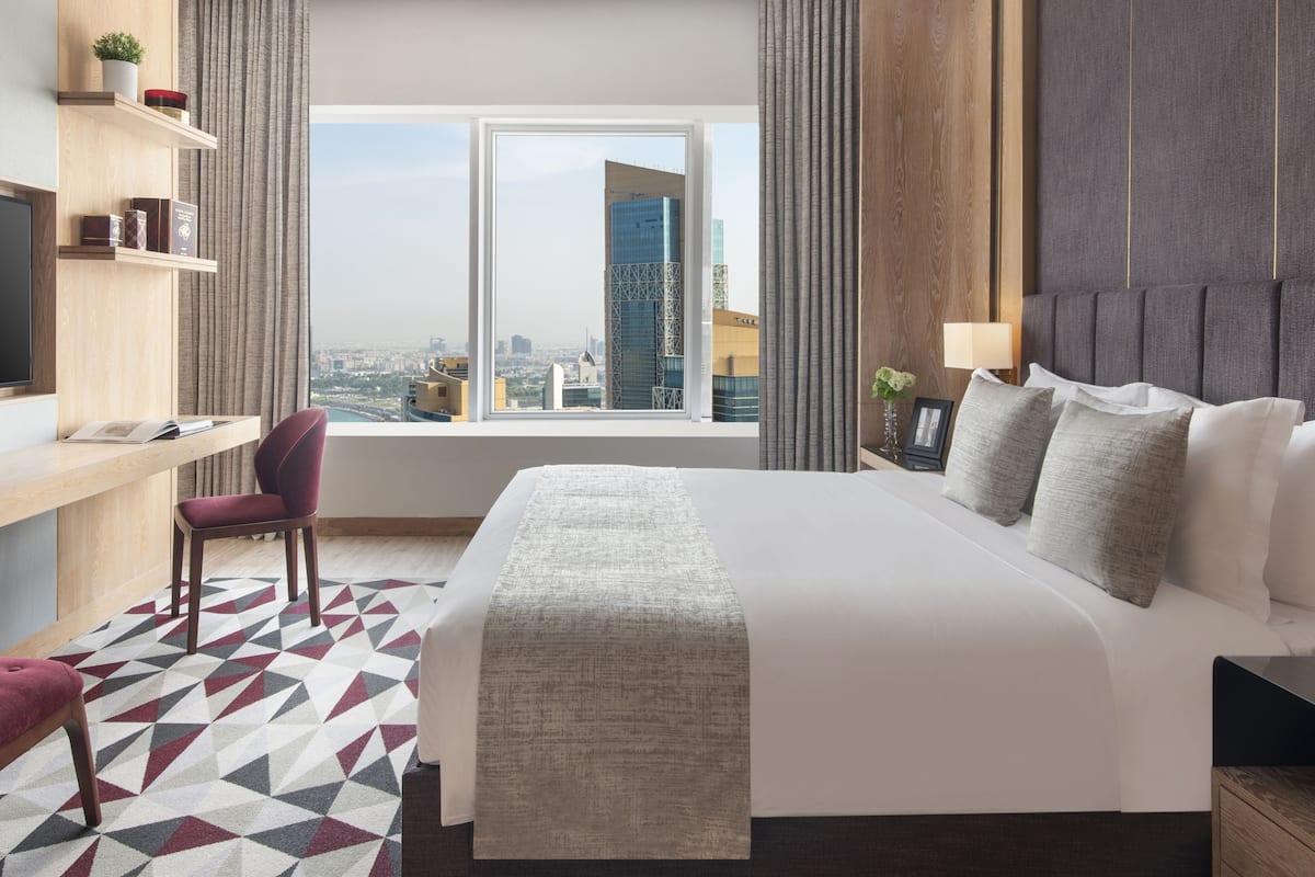 Una habitación de hotel moderna con una cama grande, un escritorio, una silla y vistas a un paisaje urbano a través de un ventanal amplio.