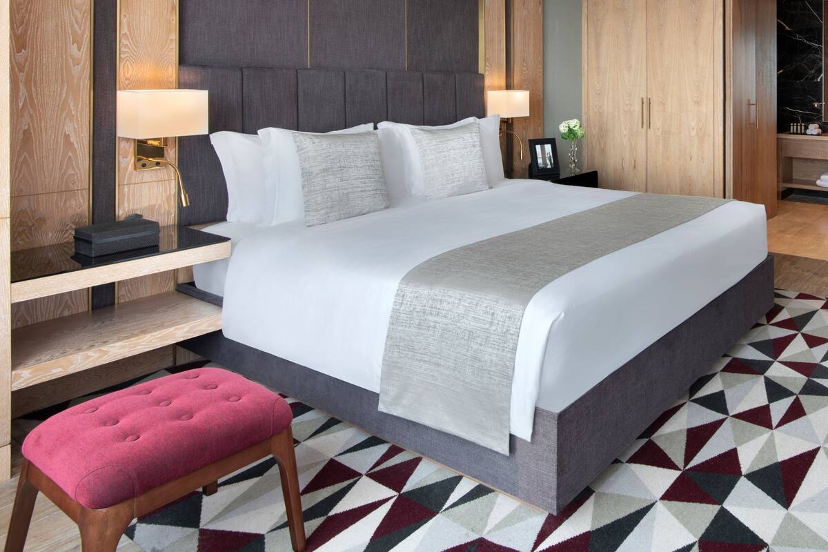 Una habitación de hotel moderna con una cama grande, lámparas de noche, un banco de madera y una alfombra con estampado geométrico.