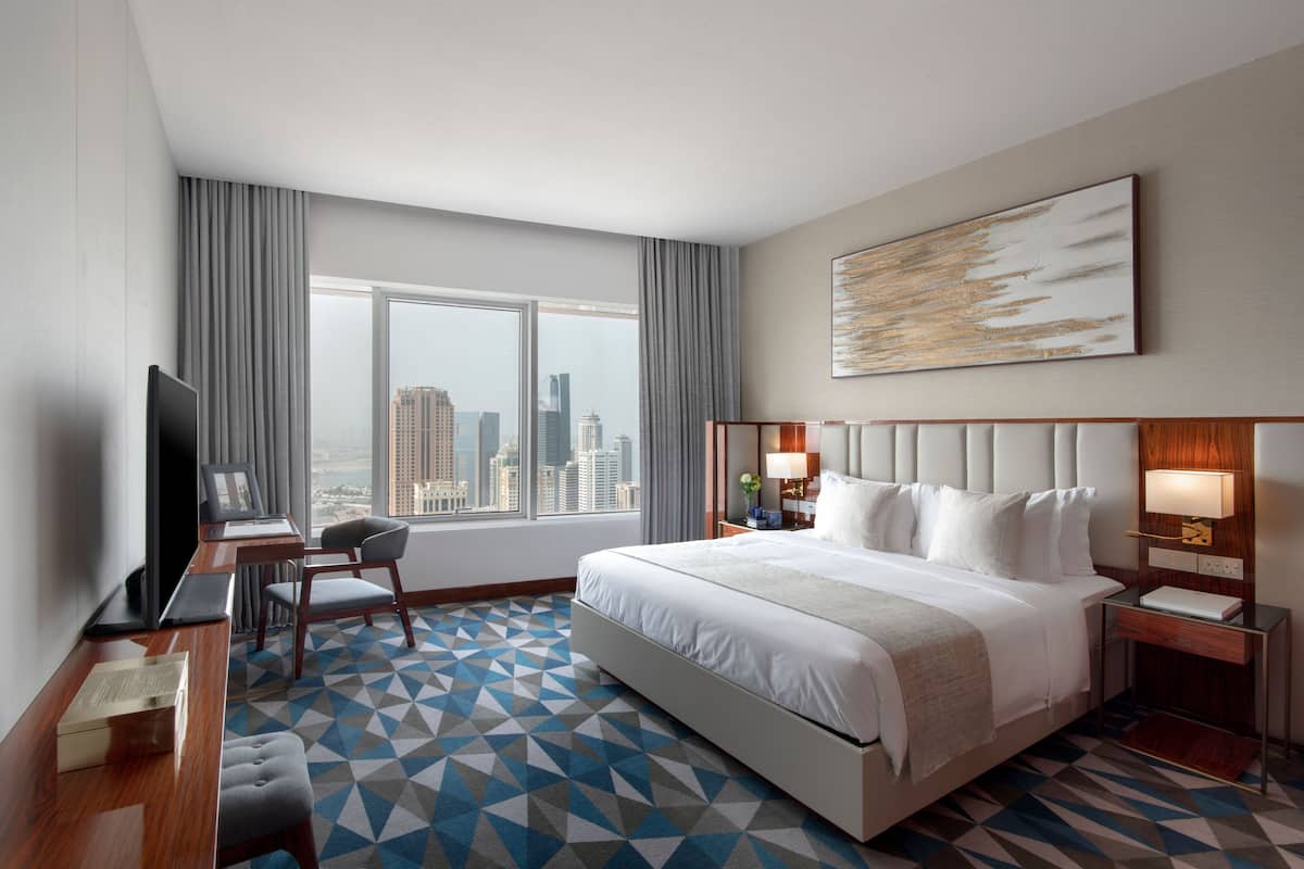 Habitación de hotel con una cama grande, un escritorio con computadora, televisor y vista a la ciudad.