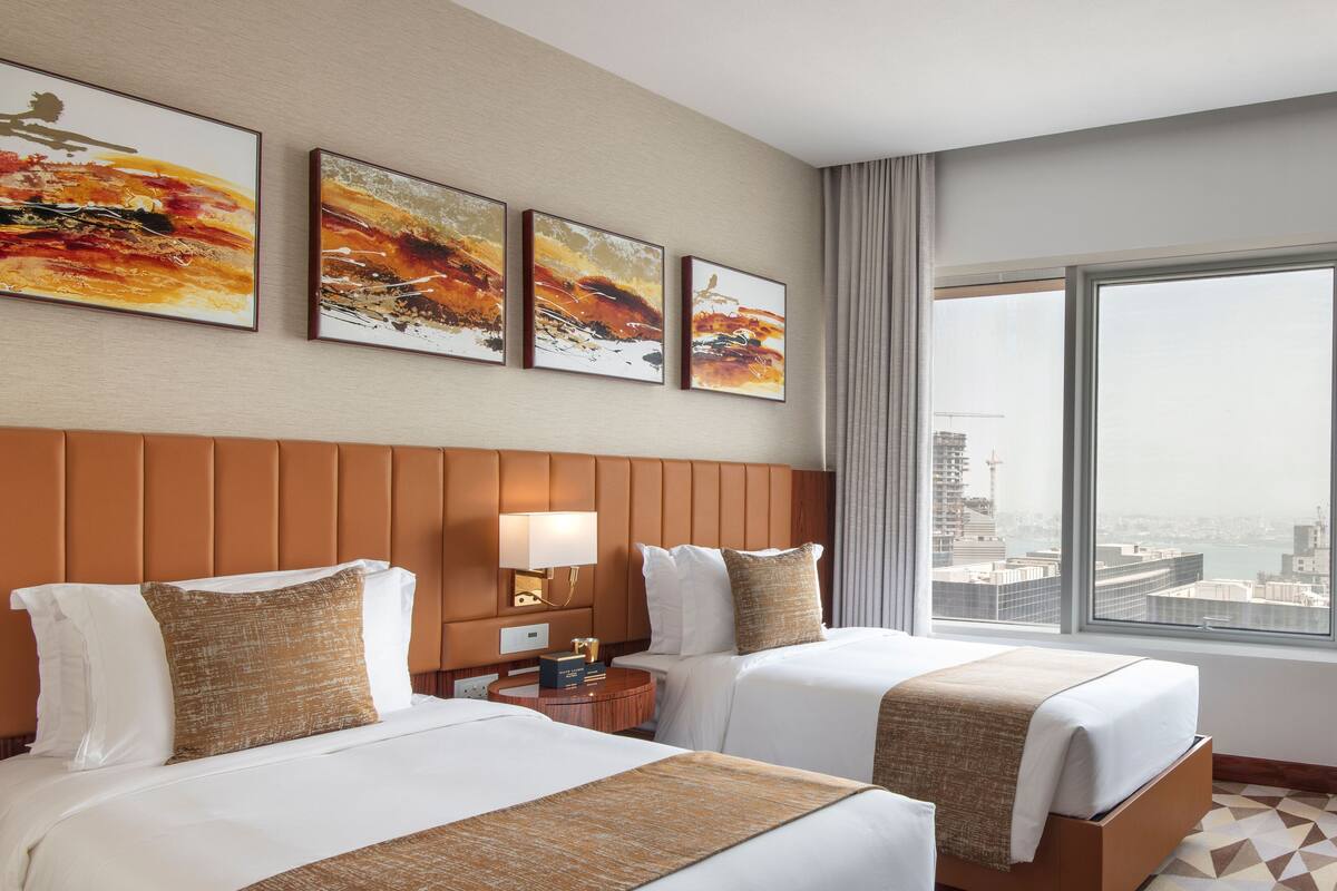 Habitación de hotel con dos camas, un ventanal amplio con vista a la ciudad y tres cuadros abstractos en la pared.