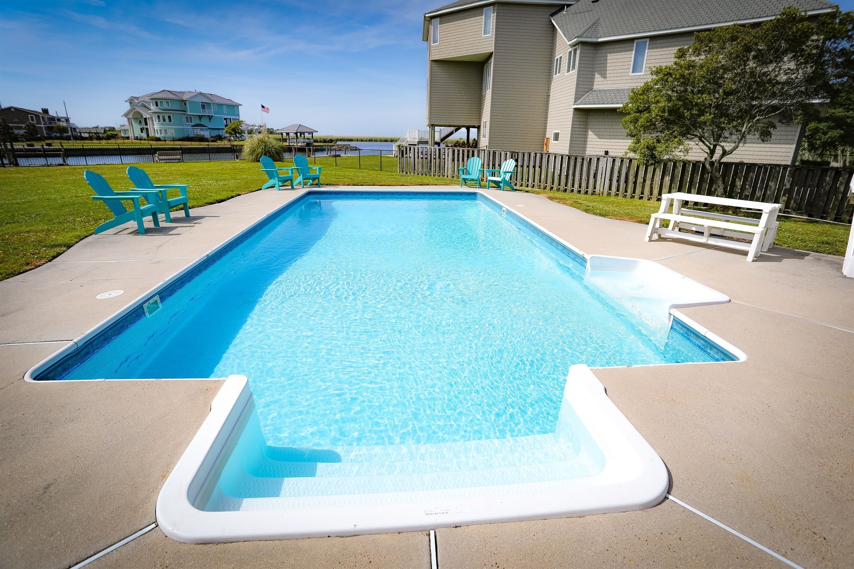A rectangular swimming pool with a white edge, surrounded by a concrete deck. There are two blue Adirondack chairs and a white bench on the deck.