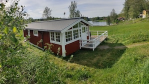 A red and white wooden cabin with a balcony, surrounded by lush greenery and a lake. - Charming 2-bed House at the Lake Close Gustavsfors (Gustavsfors)